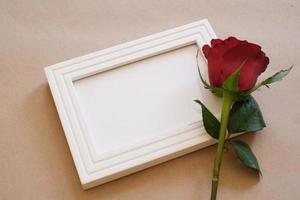 Top view of red rose laying on a white blank photo frame isolated on brown background. Valentine's day, wedding, birthday and special occasion concept. Flat lay, top view.