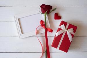 Red rose with red ribbon on a white blank photo frame and red gift box on white wooden background. Valentine's day, birthday and special occasion concept. Flat lay, top view.