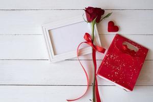 Red rose with red ribbon on a white blank photo frame and red gift bag on white wooden background. Valentine's day, birthday and special occasion concept. Flat lay, top view.