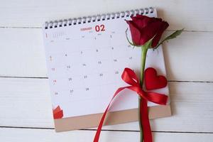 Red rose and a heart on calendar isolated on white wooden background with. Valentine's day concept. Planning scheduling agenda, Event, organiser valentines day. Flat lay, top view. photo
