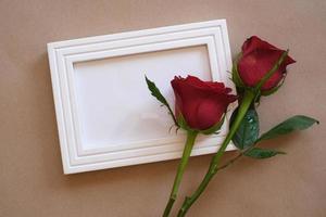 Top view of red rose and red heart laying on a white blank photo frame isolated on brown background. Valentine's day, wedding, birthday and special occasion concept. Flat lay, top view.
