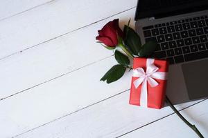 rosa roja y caja de regalo roja con portátil sobre fondo blanco de madera con. concepto de día de san valentín, boda, cumpleaños y ocasiones especiales. copie el espacio para el texto. vista superior, endecha plana. foto