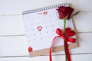 Red rose and a heart on calendar isolated on white wooden background with. Valentine's day concept. Planning scheduling agenda, Event, organiser valentines day. Flat lay, top view. photo