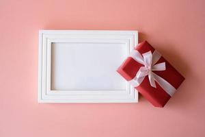 Red gift box and empty photo frame isolated on pink background for giving in holidays with copy space for text. Holidays, present, giving. New year day, Christmas day, Chinese New Year day, Birthday.