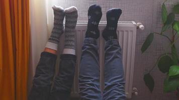 Couple hold legs up heating feet in cold home indoors on radiator in winter with cozy winter stylish woolen socks on. Valentines funny together warm feet by radiator by window video