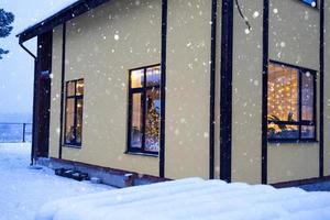 acogedora ventana festiva de la casa exterior con la cálida luz de las guirnaldas de luces de hadas en el interior: celebre la navidad y el año nuevo en un hogar cálido. árbol de navidad, bokeh, nieve en pinos y nevadas foto