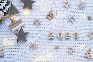 Happy New Year-wooden letters and the numbers 2023 on festive white knitted background with sequins, stars, lights of garlands. Greetings, postcard. Calendar, cover photo