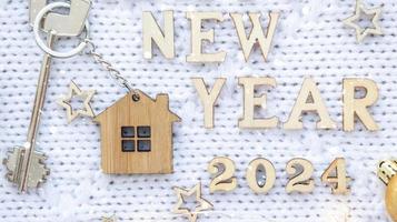 House key with keychain cottage on festive knitted background with stars, lights of garlands. Happy New Year 2024-wooden letters, greeting card. Purchase, construction, relocation, mortgage photo