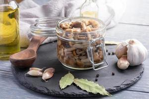 Homemade pickled honey agarics in open glass jar, spices, spoon, napkin, oil bottles on grey wooden table. Close-up. photo