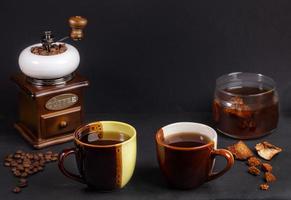 preparación café de hongos chaga. dos tazas de cerámica de dos colores, frasco de vidrio con bebida chaga, molinillo de café en negro. foto