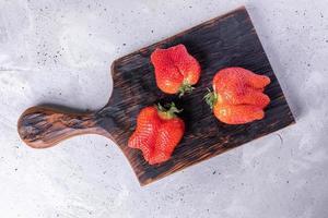 Three ugly ripe red strawberries on burned kitchen cutting board on grey concrete background. photo