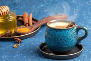 Cup of traditional Indian hot drink golden turmeric milk with cinnamon, turmeric, anise and honey on blue background.