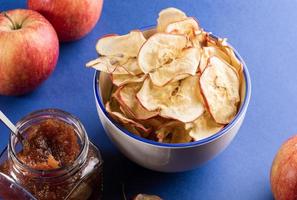 taza de cerámica blanca con patatas fritas de manzana, manzanas rojas frescas y tarro de cristal de mermelada de manzana sobre fondo azul. foto