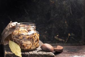 frasco de vidrio con agáricos de miel de champiñones en escabeche y cuchara de madera sobre una tabla de madera vieja sobre un fondo de metal oxidado oscuro. foto