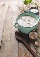 sartén de cerámica turquesa con sopa de crema de champiñones, taza con crema agria, tazón con champiñones frescos, cuchara, ajo sobre arpillera sobre fondo de madera. foto