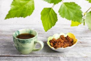 Green cup of healing beverage of birch mushroom chaga, bowl with chaga pieces on wooden table and green birch leaves. photo