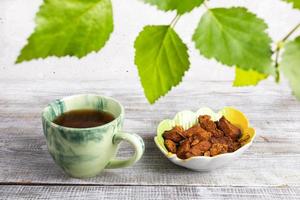 taza verde de bebida curativa de chaga de hongos de abedul, cuenco con piezas de chaga sobre mesa de madera y hojas de abedul verde. foto