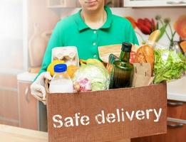 Young woman in green shirt and protective gloves holding box with food products and inscription Safe delivery. photo