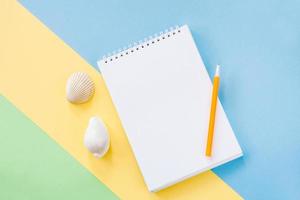 Summer vacations concept with yellow stripe, white notepad, orange pen and two seashells on blue-green background. photo