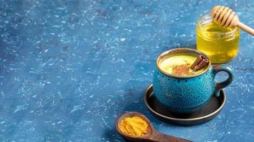 Cup of golden turmeric milk, wooden spoon with curcuma powder and jar of honey on blue. Selective focus. Copy space. photo