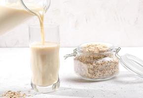 Oat milk pouring from  glass decanter into drinking glass and glass jar with oat flakes on light background. photo