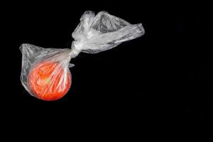 One red tomato in transparent closed plastic bag on black background. photo