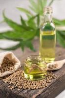 Hemp oil in two glass jar and bottle with grain in the sack on wooden board with leaves of cannabis on background.