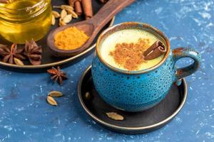 Blue cup of traditional indian golden turmeric latte milk with cardamom, anise, honey and cinnamon stick on blue. photo