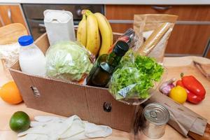 Fresh delivered food products in cardboard box and on wooden table and protective gloves. Safe delivery. Food donation. Selective focus. photo