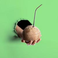 Female hand holding out one fresh coconut with metal straw inserted in it through torn hole in neo mint colored paper. photo