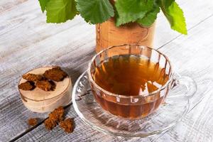 Glass cup of healing infusion of birch mushroom chaga, stump with chaga pieces and leaves on wooden table closeup. photo
