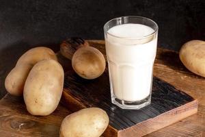 Glass of organic milk from potato and potato tubers on wooden serving board on black background.
