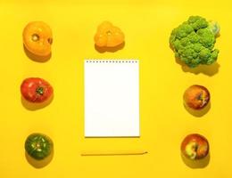 Frame of vegetables and fruits around empty clean white notepad on bright yellow background with copy space for text. photo