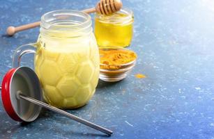 Mason jar of golden turmeric milk with metal straw, curcuma powder and honey jar on blue. Selective focus, copy space. photo