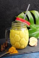Mason jar of golden turmeric milk with metal straw, ingredients for its cooking, monstera leaf on black. Vertical photo. photo