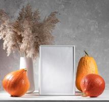 maqueta festiva de otoño con calabazas, marco vacío, hierba seca sobre una mesa blanca sobre fondo de pared de hormigón. foto