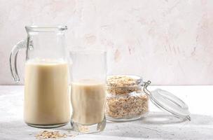 Transparent decanter and glass of oat milk and jar with oat flakes on white background. photo