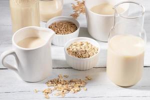 Close-up glass bottle and mug with oat milk and bowls with oat seeds and flakes on white wooden table. photo