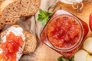 Close-up open glass jar  full of apple jam, sliced  bread, smeared with curd and apple jam on wooden table. photo