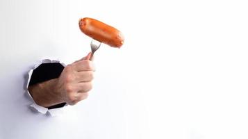 Man's hand holding hot boiled wiener on fork through ripped hole in white paper wall.