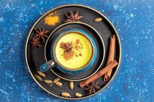 Top view of plates with cup of traditional Indian ayurvedic golden turmeric milk and ingredients on blue background. photo