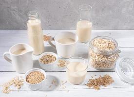 Set of different containers with oat milk, oat seeds and flakes on white wooden table on concrete background. photo