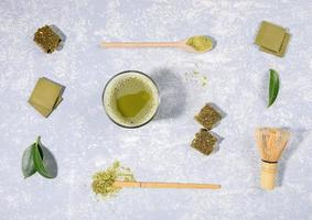 Flat lay arrangement of matcha tea glass, wooden spoons with matcha powder, whisk, leaves, chocolate, granola bar on light grey background photo