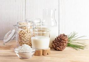 Glass and bottle with cedar milk, flour in bowl and nuts in glass jar on wooden table. Milk replacer photo