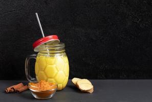 Mason jar of golden turmeric milk with metal straw, ingredients for its cooking on black background. Copy space. photo