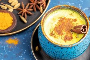 Close-up cup of traditional indian ayurvedic golden turmeric latte milk with cardamom, anise and cinnamon on blue. photo