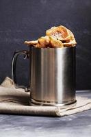 Metal mug with apple chips standing on burlap on dark background.