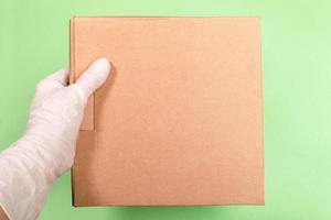 Female hand in white latex glove holding brown cardboard box on light green backdrop. Concept safe delivery. Copy space. photo