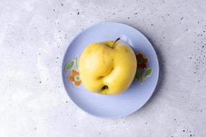 una fea manzana amarilla sobre un plato lila sobre fondo de hormigón gris. foto