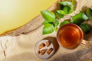 Glass cup of healing drink of birch mushroom chaga, mushroom pieces, twig on wooden plank on yellow backdrop. Top view. photo